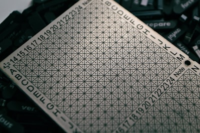 A metallic board with a grid of small triangular patterns, surrounded by letters and numbers. The board is placed over scattered dark tiles with engraved words that are partially visible.