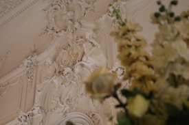 An ornate and intricate plaster ceiling is decorated with detailed carvings and sculptural elements, featuring floral and foliage motifs. In the foreground, blurred yellow flowers add depth to the image.