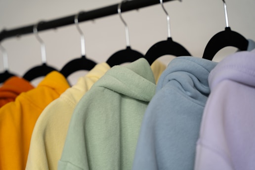 A row of colorful hoodies displayed on black hangers. The hoodies are in shades of orange, yellow, green, blue, and lavender, arranged in a gradient order.