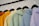 A row of colorful hoodies displayed on black hangers. The hoodies are in shades of orange, yellow, green, blue, and lavender, arranged in a gradient order.