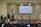 A speaker stands at a podium on a stage, with two flags on either side. A large screen behind displays 'DEFENSOR TEC'. Two empty chairs and a small round table are also on the stage. Audience members are visible in the foreground.