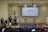 A speaker stands at a podium on a stage, with two flags on either side. A large screen behind displays 'DEFENSOR TEC'. Two empty chairs and a small round table are also on the stage. Audience members are visible in the foreground.