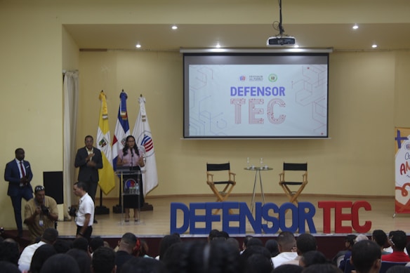 A speaker stands at a podium on a stage, with two flags on either side. A large screen behind displays 'DEFENSOR TEC'. Two empty chairs and a small round table are also on the stage. Audience members are visible in the foreground.