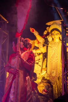 A traditional festival scene featuring a woman in ornate attire holding a decorated staff, with an elaborate idol of a deity in the background. The image is rich in cultural symbolism and vibrant colors.