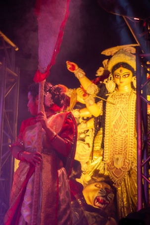 A traditional festival scene featuring a woman in ornate attire holding a decorated staff, with an elaborate idol of a deity in the background. The image is rich in cultural symbolism and vibrant colors.