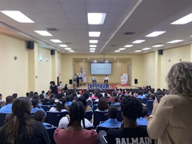 A large group of people seated in a modern auditorium, attentively facing a stage where a speaker is presenting. Several banners and flags are visible on the stage, along with a projection screen displaying some logos. The room is brightly lit with ceiling lights, and there are various attendees of different ages and backgrounds, some taking notes or photos.