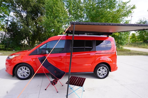 A bright red van parked on a concrete surface with an extended black awning. In front of the van, there is a folding chair and a small portable table on the ground. The background consists of lush green trees under a clear blue sky.