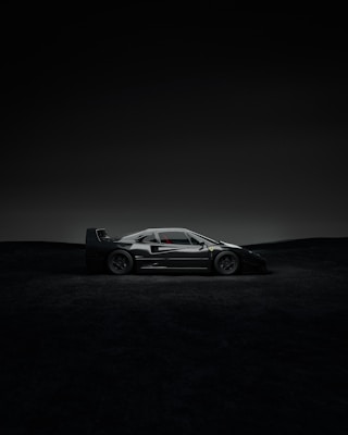 A sleek, dark sports car is parked in a dramatic setting with minimal lighting. The car's design is aerodynamic, with a low profile and sharp angles, highlighted subtly by the dim environment. The background features a nearly black landscape that enhances the focus on the vehicle.