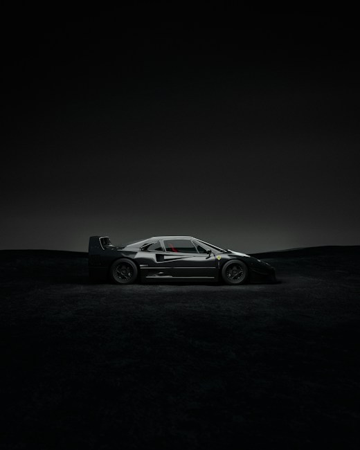 A sleek, dark sports car is parked in a dramatic setting with minimal lighting. The car's design is aerodynamic, with a low profile and sharp angles, highlighted subtly by the dim environment. The background features a nearly black landscape that enhances the focus on the vehicle.