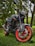 A sleek, black motorbike with red highlights on its rims and trim, parked on a grassy area surrounded by trees and foliage. The bike is detailed with modern components and appears well-maintained.