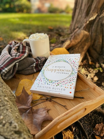 A cozy autumn scene with a wooden tray on a bed of fallen leaves. A mug of frothy hot beverage sits beside a plaid scarf, next to a book titled '3 Minute Happiness Journal' by Kristen Butler. The background features a large tree trunk and blurred greenery.