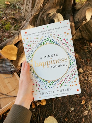 A hand holds a journal titled '3 Minute Happiness Journal' by Kristen Butler. The cover features colorful dot patterns, and the phrase 'Create happy habits. Change your brain. Transform your life.' is visible at the top. The background includes a tree trunk and fallen leaves.