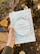 A hand holds a journal titled '3 Minute Happiness Journal' by Kristen Butler. The cover features colorful dot patterns, and the phrase 'Create happy habits. Change your brain. Transform your life.' is visible at the top. The background includes a tree trunk and fallen leaves.