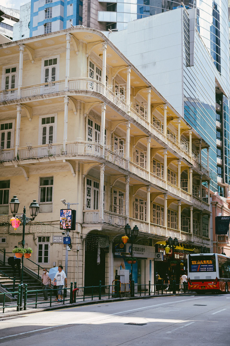 The image depicts a street scene with a tall, two-story building painted white and adorned with balconies on each floor. The building features a curved corner at the top, adding architectural interest to its design. A bus is parked in front of the building, suggesting it may be a popular transportation hub or commercial area.

On the sidewalk next to the building, there are two people walking, one wearing a white shirt and carrying a backpack. The street itself appears to be relatively empty, with no other vehicles visible.