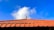 A traditional terracotta roof is visible against a vibrant blue sky with a few fluffy white clouds. The roof has a distinct pattern with uniformly arranged tiles and a decorative finial at the top center.