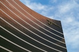 A tall, modern building with reflective glass windows curves gracefully against a backdrop of a partly cloudy blue sky. The building features a distinctive logo at the top, adding a touch of elegance.
