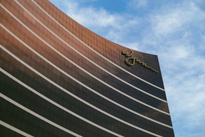 A tall, modern building with reflective glass windows curves gracefully against a backdrop of a partly cloudy blue sky. The building features a distinctive logo at the top, adding a touch of elegance.