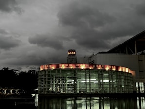A modern, circular glass building is set against a backdrop of dark, overcast skies. The structure features a red illuminated sign with various phrases such as 'Knowledge' and 'International Standards' in English and another language. Reflections from the building appear on the water in the foreground, enhancing the moody atmosphere.