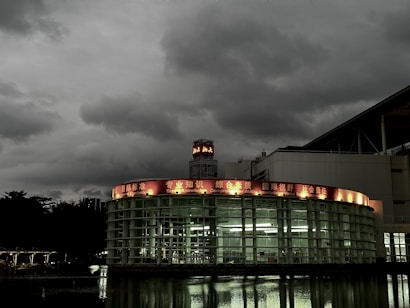 A modern, circular glass building is set against a backdrop of dark, overcast skies. The structure features a red illuminated sign with various phrases such as 'Knowledge' and 'International Standards' in English and another language. Reflections from the building appear on the water in the foreground, enhancing the moody atmosphere.