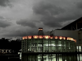 A modern, circular glass building is set against a backdrop of dark, overcast skies. The structure features a red illuminated sign with various phrases such as 'Knowledge' and 'International Standards' in English and another language. Reflections from the building appear on the water in the foreground, enhancing the moody atmosphere.