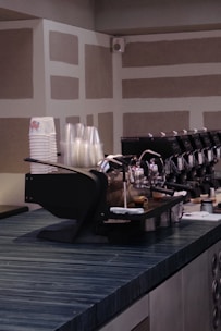 A modern coffee counter with a sleek espresso machine. The countertop is dark and textured, with a stack of paper and plastic cups on the side. The background features a geometric pattern on the walls.