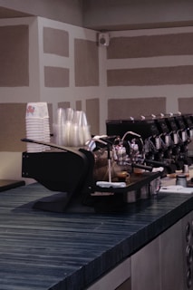 A modern coffee counter with a sleek espresso machine. The countertop is dark and textured, with a stack of paper and plastic cups on the side. The background features a geometric pattern on the walls.