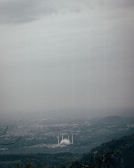 A vast, cloudy skyline overlooks a sprawling cityscape with a prominent white mosque featuring minarets visible in the center. The landscape includes lush greenery and hills in the foreground, while the city extends into the distance under heavy clouds.