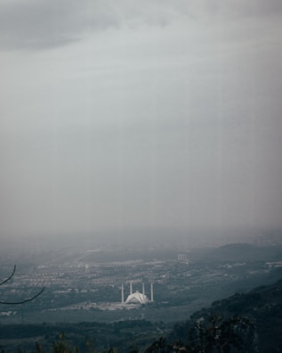 A vast, cloudy skyline overlooks a sprawling cityscape with a prominent white mosque featuring minarets visible in the center. The landscape includes lush greenery and hills in the foreground, while the city extends into the distance under heavy clouds.