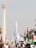 A large group of people gathered at a protest rally. Many are holding flags, including Palestinian flags, and signs that advocate for boycotting Israel. The event is taking place outdoors near a tall monument with a flame structure on top. Palm trees and city buildings can be seen in the background.