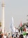 A large group of people gathered at a protest rally. Many are holding flags, including Palestinian flags, and signs that advocate for boycotting Israel. The event is taking place outdoors near a tall monument with a flame structure on top. Palm trees and city buildings can be seen in the background.
