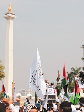 A large group of people gathered at a protest rally. Many are holding flags, including Palestinian flags, and signs that advocate for boycotting Israel. The event is taking place outdoors near a tall monument with a flame structure on top. Palm trees and city buildings can be seen in the background.