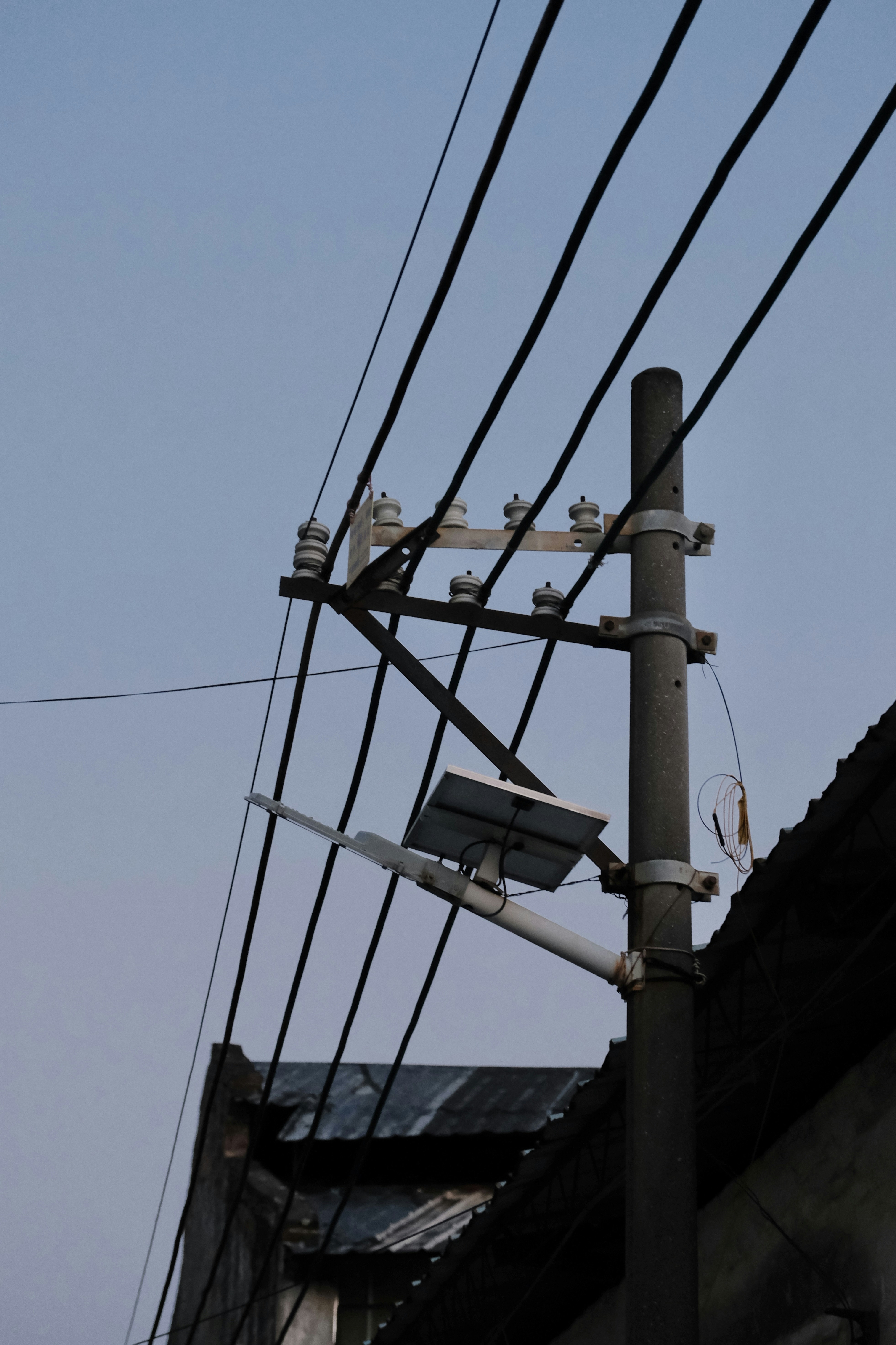 A street light pole with electrical wires stretching across it, along with a mounted solar panel near the top. The background shows the edge of a corrugated roof, under a clear sky.
