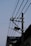 A street light pole with electrical wires stretching across it, along with a mounted solar panel near the top. The background shows the edge of a corrugated roof, under a clear sky.