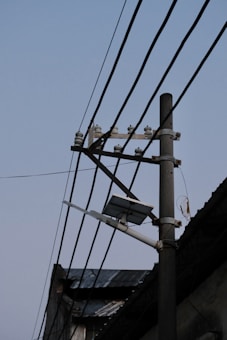A street light pole with electrical wires stretching across it, along with a mounted solar panel near the top. The background shows the edge of a corrugated roof, under a clear sky.