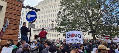 A large crowd gathers in a city area, holding signs and banners advocating for Gaza. The atmosphere is busy with people standing, walking, and actively engaging in the demonstration. Buildings and trees are visible in the background, along with street signs.
