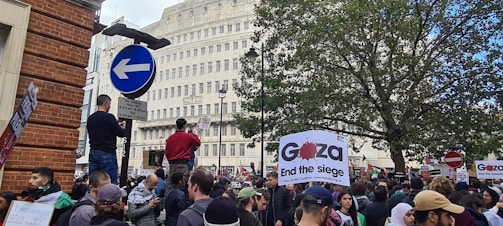 A large crowd gathers in a city area, holding signs and banners advocating for Gaza. The atmosphere is busy with people standing, walking, and actively engaging in the demonstration. Buildings and trees are visible in the background, along with street signs.