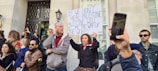 A group of people participate in a protest or demonstration outside a building with ornate iron doors. The central figure holds a sign that reads, 'If we don't heal Palestine, Palestine will become the way of the world.' Several individuals use their phones to take photos or record the event.