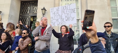 A group of people participate in a protest or demonstration outside a building with ornate iron doors. The central figure holds a sign that reads, 'If we don't heal Palestine, Palestine will become the way of the world.' Several individuals use their phones to take photos or record the event.