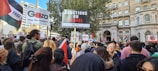 A large group of people gathered in a city environment holding signs and flags. Prominent signs read 'Sanctions on Israel', 'Gaza stop the massacre', and 'Free Palestine'. The crowd includes diverse individuals, and the scene suggests a protest or demonstration. Surrounding buildings include classic architectural elements.