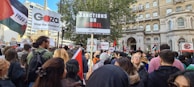 A large group of people gathered in a city environment holding signs and flags. Prominent signs read 'Sanctions on Israel', 'Gaza stop the massacre', and 'Free Palestine'. The crowd includes diverse individuals, and the scene suggests a protest or demonstration. Surrounding buildings include classic architectural elements.