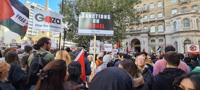 A large group of people gathered in a city environment holding signs and flags. Prominent signs read 'Sanctions on Israel', 'Gaza stop the massacre', and 'Free Palestine'. The crowd includes diverse individuals, and the scene suggests a protest or demonstration. Surrounding buildings include classic architectural elements.
