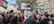 A large group of people gathered in a city environment holding signs and flags. Prominent signs read 'Sanctions on Israel', 'Gaza stop the massacre', and 'Free Palestine'. The crowd includes diverse individuals, and the scene suggests a protest or demonstration. Surrounding buildings include classic architectural elements.