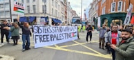 A group of people are participating in a street protest. They hold a large banner that reads 'Sisters Stand with Gaza Free Palestine' and smaller placards with messages supporting Palestine. The setting is an urban street lined with buildings, and there is a truck and some construction activity in the background.