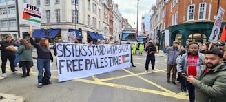 A group of people are participating in a street protest. They hold a large banner that reads 'Sisters Stand with Gaza Free Palestine' and smaller placards with messages supporting Palestine. The setting is an urban street lined with buildings, and there is a truck and some construction activity in the background.