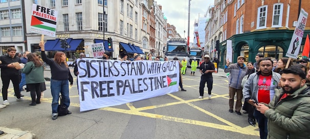 A group of people are participating in a street protest. They hold a large banner that reads 'Sisters Stand with Gaza Free Palestine' and smaller placards with messages supporting Palestine. The setting is an urban street lined with buildings, and there is a truck and some construction activity in the background.