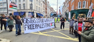 A group of people are participating in a street protest. They hold a large banner that reads 'Sisters Stand with Gaza Free Palestine' and smaller placards with messages supporting Palestine. The setting is an urban street lined with buildings, and there is a truck and some construction activity in the background.