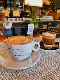 A beautifully presented cappuccino in a decorative cup sits on a woven placemat, accompanied by a glass of layered espresso on a black saucer. The background features a cozy café ambiance with warm lighting, potted plants, and a QR code on a menu stand.