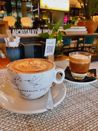 A beautifully presented cappuccino in a decorative cup sits on a woven placemat, accompanied by a glass of layered espresso on a black saucer. The background features a cozy caf&eacute; ambiance with warm lighting, potted plants, and a QR code on a menu stand.