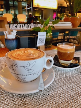 A beautifully presented cappuccino in a decorative cup sits on a woven placemat, accompanied by a glass of layered espresso on a black saucer. The background features a cozy caf&eacute; ambiance with warm lighting, potted plants, and a QR code on a menu stand.