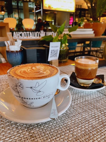 A beautifully presented cappuccino in a decorative cup sits on a woven placemat, accompanied by a glass of layered espresso on a black saucer. The background features a cozy caf&eacute; ambiance with warm lighting, potted plants, and a QR code on a menu stand.